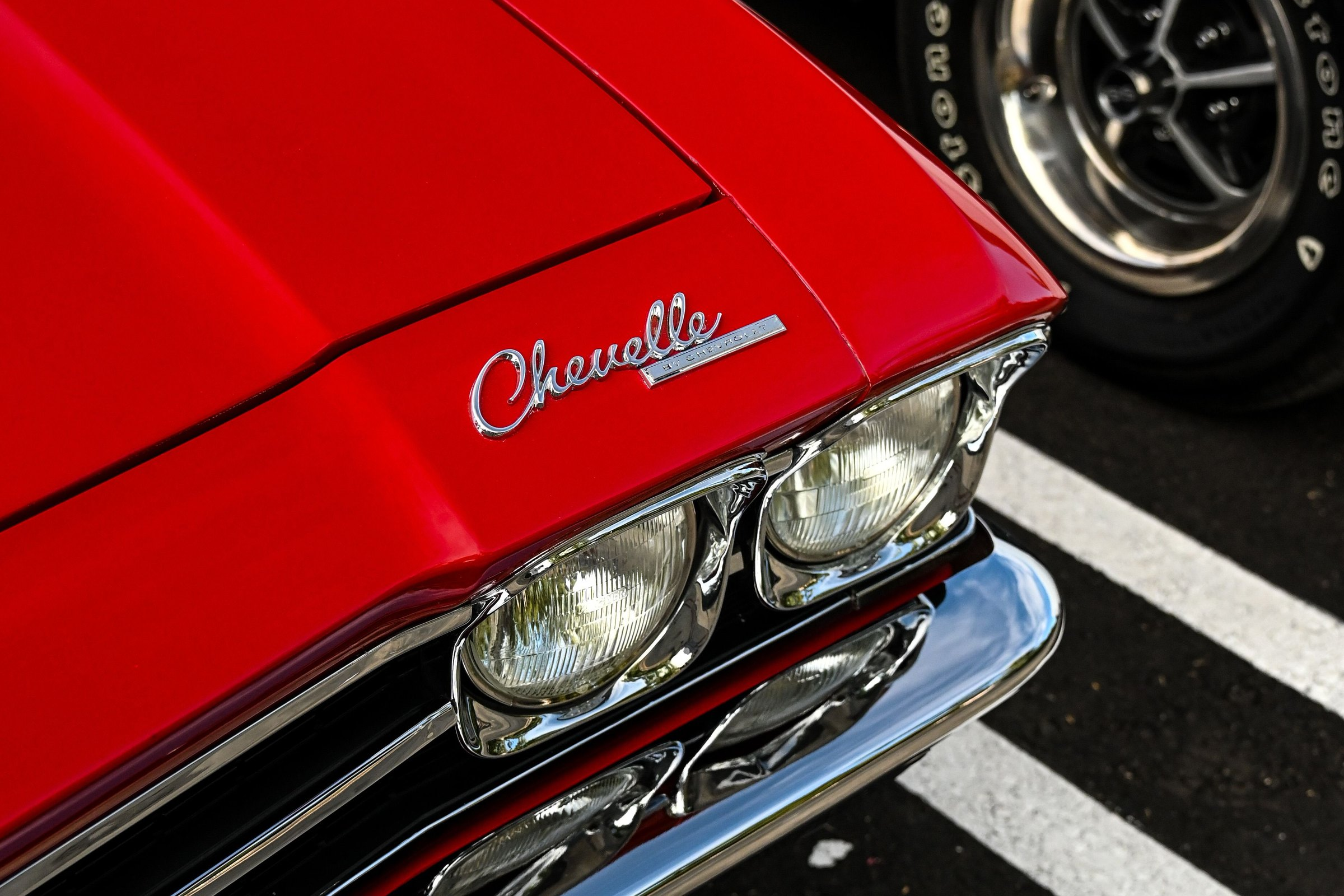 Front corner of a red classic Chevrolet Chevelle with chrome Chevelle script, headlights, and front bumper